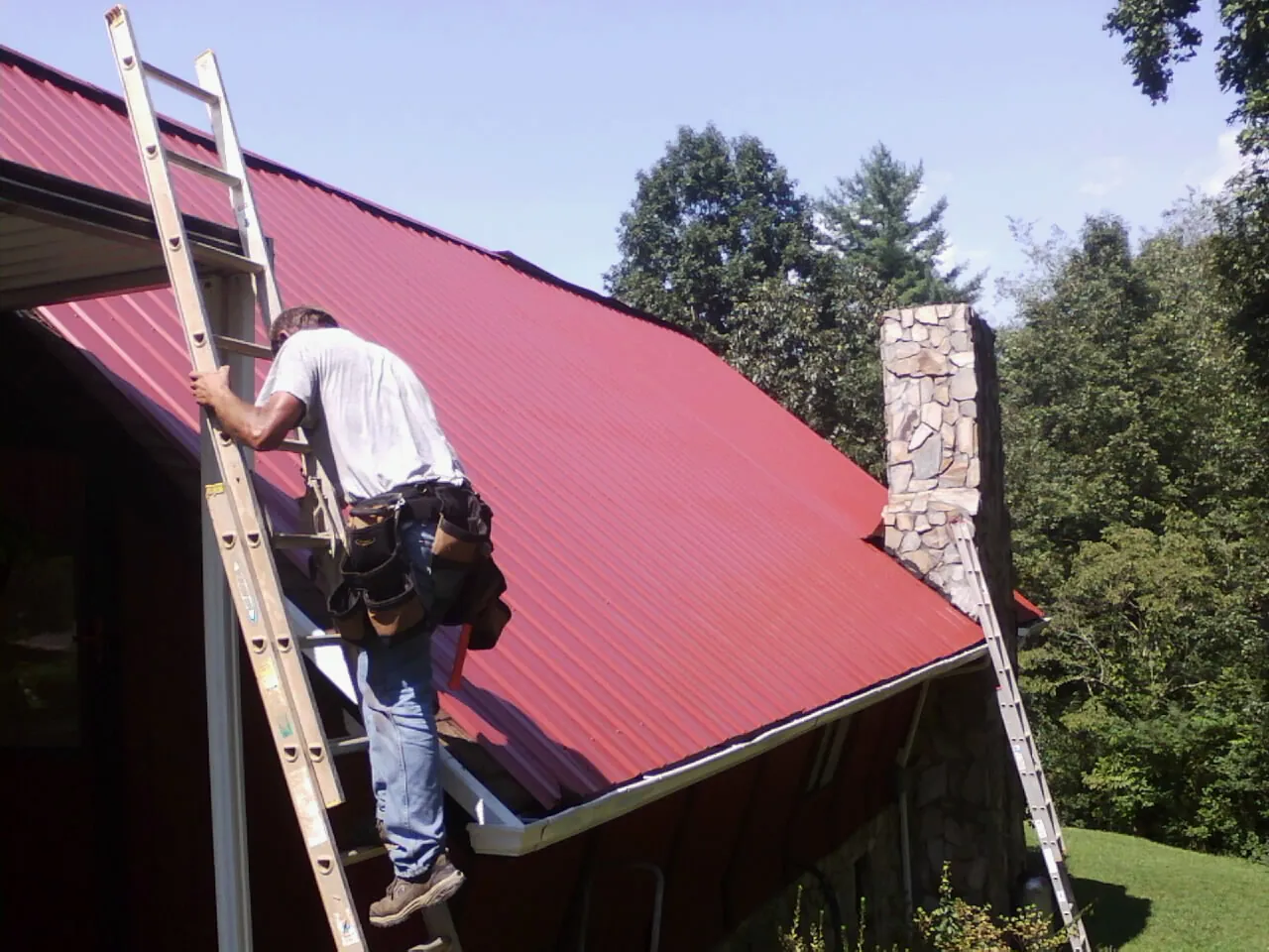 Roofer on a ladder beside a red metal roof and stone chimney — Soffit & Fascia Repair by Roofing Brothers in Fresh Meadows